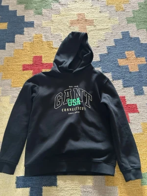 Svart hoodie från GANT med tryck - Svart hoodie från GANT med stort vitt och grönt tryck på bröstet där det står 'GANT USA Connecticut'. Hoodien har huva och ribbade muddar vid ärmslut och nederkant. Perfekt för dig som gillar klassisk college-stil.