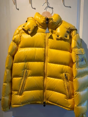 Gul dunjacka från Moncler - Snygg gul dunjacka från Moncler med hög krage, två dragkedjefickor fram och en ficka med lock och logotyp på ärmen. Jackan har quiltad design och blanka detaljer. Perfekt för kalla vinterdagar och riktigt trendig färg.