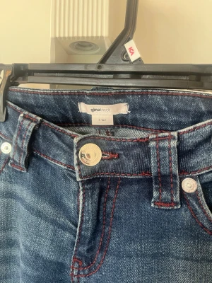 Mörkblå jeans från Gina Tricot - Snygga mörkblå jeans från Gina Tricot med röda kontrastsömmar och klassisk femficksmodell. Jeansen har en lätt tvättad look och silverfärgade knappar. Perfekta för en trendig och avslappnad stil. Sprättat upp ner till på jeansen för längre längd 