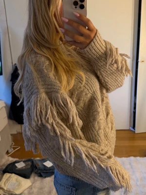 Beige stickad tröja med fransar - Säljer en beige stickad tröja med grovt kabelstickat mönster och coola fransdetaljer längs ärmarna. Tröjan har rund halsringning och ribbade muddar.
