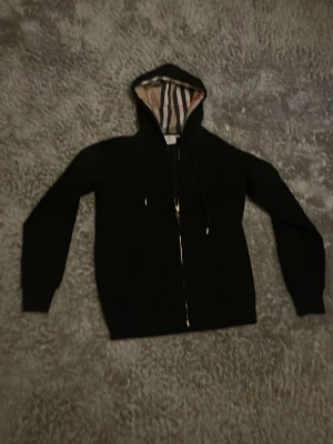 Svart hoodie från Burberry - Säljer en svart hoodie från Burberry med dragkedja och klassiskt rutigt foder i huvan. Hoodien har dragsko i huvan och är tillverkad i mjukt material. Perfekt för dig som vill ha en stilren och exklusiv look med en touch av Burberrys ikoniska design.
