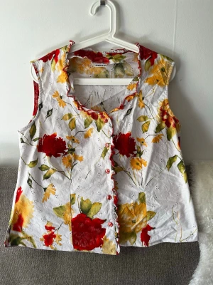 Blommig ärmlös blus med volangkant - Säljer en somrig ärmlös blus från Janina med färgglatt blommönster i rött, gult och grönt på vit botten. Blusen har volangkant längs knäppningen och en mjuk V-ringning. Perfekt för dig som gillar färg och vill sticka ut lite extra.