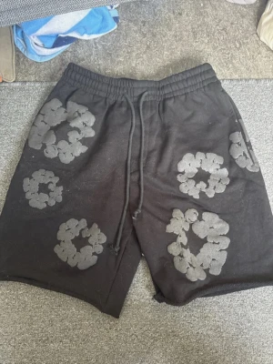 Svarta shorts med blommigt mönster - Svarta shorts med elastisk midja och snörning. Dekorerade med stora blommiga mönster i en mörkare nyans. Perfekta för en avslappnad och trendig stil. Mjukt material som känns skönt mot huden.
