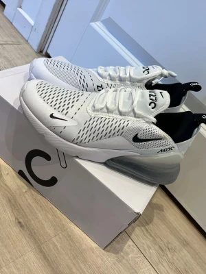 Nike Air Max 270 vita sneakers - Nike Air Max 270 sneakers i vitt med svarta detaljer och stor synlig Air-enhet i hälen. Ovandelen är i mesh för bra ventilation och har en modern, sportig look. Klassisk Nike-logga på sidan och tungan. Skön passform med vadderad häl och snörning.