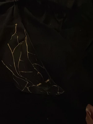 Svart liten väska med mönster - Säljer en svart liten väska med dragkedja och abstrakt mönster i beige. Perfekt att ha småsaker i och lätt att ta med sig. Väskan har en mjuk form och är tillverkad i tygmaterial.