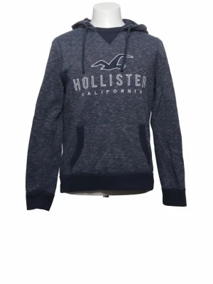 Mörkblå hoodie från Hollister - Mörkblå hoodie från Hollister med vit logga och texten 'California' på bröstet. Hoodien har huva med dragsko, magficka och ribbade muddar vid ärmslut och nederkant. Tillverkad i mjukt sweatshirtmaterial som är perfekt för chill dagar.