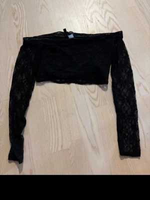 Svart offshoulder spets topp - Trendig svart offshoulder topp i spets med långärmad design. Croppad modell som passar perfekt till höga jeans eller kjol. Tunn och lätt, perfekt för att skapa en edgy look.