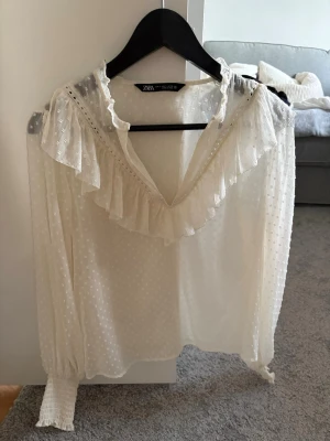 Vit volangblus från Zara - Vit blus från Zara med volangdetaljer längs v-ringningen och axlarna. Blusen har diskret prickigt mönster i tyget och långa ärmar med breda manschetter. Perfekt för en trendig och feminin look.