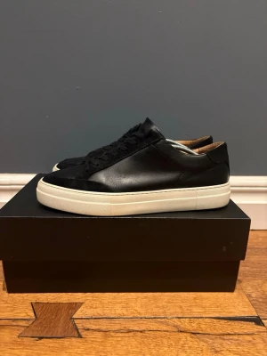 Svarta j lindberg sneakers - Stilrena svarta sneakers i läder med klassisk låg profil och tjock vit sula. Skorna har svarta skosnören och en enkel, clean design som passar till det mesta. Insidan är fodrad i brunt läder för extra komfort.