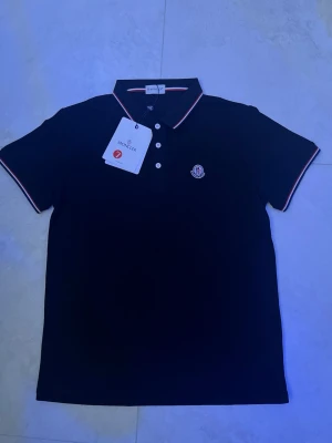 Svart pikétröja från Moncler - Klassisk svart pikétröja från Moncler med vit och röd kant på krage och ärmslut. Tröjan har korta ärmar, knappslå framtill och Moncler-logga på bröstet. Perfekt för en stilren och sportig look.