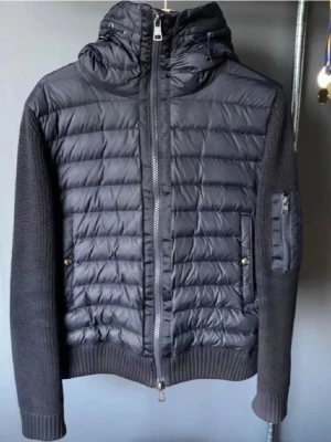 Svart moncler cardigan - Snygg svart cardigan med vadderad framsida och huva. Ärmar och rygg är stickade i grov bomull, vilket ger en cool kontrast. Jackan har dragkedja framtill, fickor med tryckknappar och en extra ficka på ärmen. Perfekt för kalla vinterdagar.