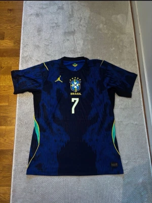 Blå Brasilien fotbollströja Vinicius Jr - Snygg blå fotbollströja från Nike med Brasilien-emblem och nummer 7 på bröstet. På ryggen står det Vinicius Jr och stort nummer 7. Tröjan har gula och gröna detaljer längs sidorna och är tillverkad i ett lätt, ventilerande material perfekt för träning eller match.
