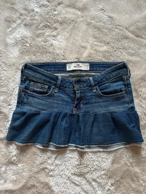 Jeanskjol med volang från Hollister - Snygg blå jeanskjol från Hollister med volang nertill och klassiska fickor fram och bak. Kjolen har en låg midja och är perfekt för dig som gillar en avslappnad och trendig stil. Ikonisk Hollister-logga på fickan och slitna detaljer för extra edge.
