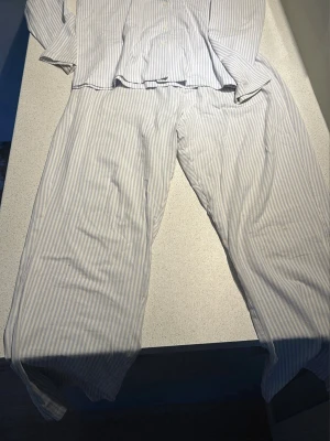 Randig pyjamas från Kappahl - Klassisk tvådelad pyjamas från Kappahl i vitt med ljusblå smala ränder. Skjortan har knappar framtill och byxorna är raka i modellen. Tillverkad i mjuk bomull, perfekt för mysiga kvällar hemma.