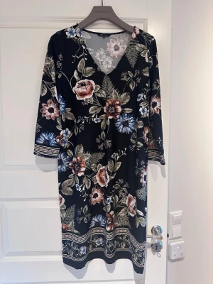Svart blommig klänning med v-ringning - Svart klänning med v-ringning och trekvartsärm, täckt av stora blommor i beige, blått och grönt. Klänningen har dekorativa bårder vid ärmslut och nederkant. Perfekt för dig som gillar en romantisk och bohemisk stil.