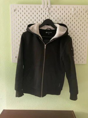 Svart hoodie med dragkedja - Säljer en svart hoodie från Mountain Warehouse med dragkedja framtill och två fickor. Insidan av huvan är fodrad med mjukt, vitt teddyfoder som gör den extra skön. Klassisk passform och ribbade muddar vid ärmslut och nederkant. Perfekt för dig som gillar enkel och stilren design. SNABB AFFÄR!