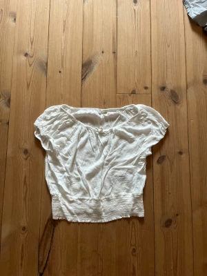 Vit croppad blus med rosett - Söt vit blus med korta ärmar och croppad passform. Blusen har en liten rosett framtill vid halsen och smockad nederkant som ger en snygg siluett. Perfekt för sommarens varma dagar och enkel att matcha med jeans eller kjol.
