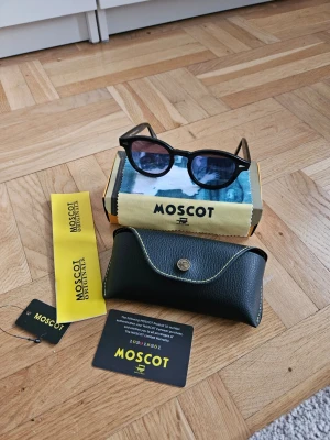 Svarta solglasögon från Moscot - Snygga svarta solglasögon från Moscot med rundade bågar och klassisk design. Tillverkade i blankt syntetmaterial och levereras med originalfodral och putsduk. Perfekta för dig som vill ha en tidlös och cool look.