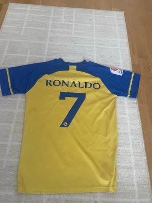 Al Nassr Ronaldo matchtröja gul/blå - Snygg Al Nassr matchtröja i gult och blått med Ronaldo och nummer 7 på ryggen. Tröjan har klubbmärke och sponsortryck på bröstet samt korta ärmar. Tillverkad i lätt och ventilerande polyester, perfekt för fotbollsträning eller att bära som supporter.