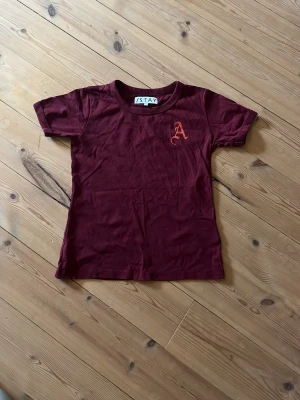 Vinröd t-shirt från ISTAY - Snygg vinröd t-shirt från ISTAY med korta ärmar och rund hals. På bröstet finns ett tryckt A i orange. Perfekt basplagg med en slim passform och mjukt bomullsmaterial.