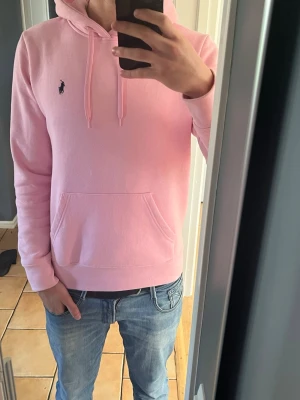 Rosa hoodie från Polo Ralph Lauren - Säljer en ljusrosa hoodie från Polo Ralph Lauren med klassisk huva och dragsko. Tröjan har en känguruficka framtill och den ikoniska mörkblå loggan broderad på bröstet. Tillverkad i mjukt bomullsmaterial som är skönt mot huden. Perfekt för en avslappnad och trendig look.