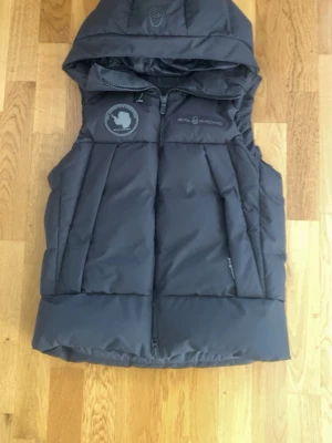 Svart dunväst med huva från Sail Racing - Cool svart dunväst från Sail Racing med huva och dragkedja framtill. Västen har ett snyggt märke på bröstet och är tillverkad i ett slitstarkt material med dunfyllning som håller dig varm. Perfekt för lager-på-lager och streetstyle.