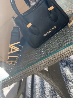 Svart handväska från Valentino - Snygg svart handväska från Valentino med guldfärgade detaljer och justerbar axelrem. Väskan har ett stilrent utseende med präglat läder och logotyp framtill. Perfekt storlek för det viktigaste och en bred axelrem med VALENTINO-text i guld. Använd 2-3 gånger               Nypris:1300kr