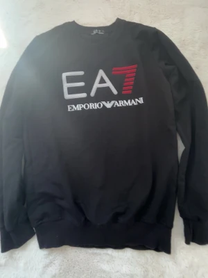 Svart sweatshirt från Emporio Armani - Snygg svart sweatshirt från Emporio Armani med stort EA7-tryck i vitt och rött på bröstet. Tröjan har rund halsringning, långa ärmar och ribbade muddar. Perfekt för en trendig och avslappnad stil.