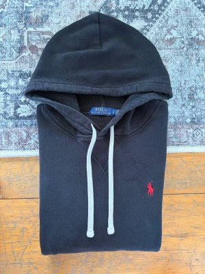 Svart Polo Ralph Lauren Hoodie - Hej! 👋 Säljer en stilren svart Polo Ralph Lauren hoodie i storlek S (Herr). Den har den klassiska röda pony-loggan broderad på bröstet 🐎 Tröjan är i bra skick och har den sköna mjuka fleece-insidan kvar ✨ Det kan förekomma en naturlig, lätt solblekning, främst på ena ärmen, vilket knappt är synligt i verkligheten 👕. Modellen saknar QR-kod men alla tvättlappar sitter kvar ✅ Ett oumbärligt basplagg som passar till allt! Fraktar snabbt 🚛📦💬💯 