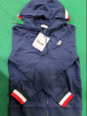 Marinblå hoodie f - Snygg marinblå hoodie från  med dragkedja, huva och ribbade muddar i rött, vitt och blått. Klassisk  på bröstet och randig detalj på huvan. Perfekt för en stilren och sportig look.