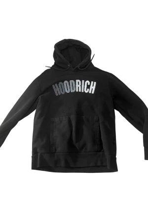 Svart hoodie från Hoodrich - Svart hoodie från Hoodrich med stor vit logga på bröstet. Klassisk känguruficka framtill och justerbar huva med snören. Tillverkad i mjukt material som känns skönt mot huden. Perfekt för en avslappnad streetwear-look.
