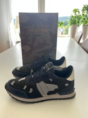 Valentino Garavani camo sneakers - Snygga sneakers från Valentino Garavani i svart och vitt med camomönster. Skorna har meshdetaljer, vita partier på hälen och en klassisk låg siluett. Perfekta för dig som vill ha en unik och trendig look. Kommer med originalkartong. Andvänd få gånger och bra kvalitet. Kom med bud!