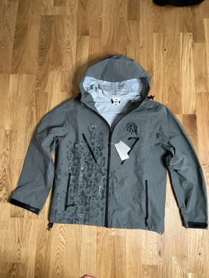 Metra windbreaker  - Snygg grå vindjacka från metra med svart grafiskt tryck på framsidan. Jackan har huva, svarta dragkedjor och två snedställda fickor framtill. Perfekt för blåsiga dagar och streetwear-stil. Materialet är lätt och vattenavvisande.