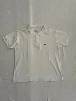 Vit pikétröja från Lacoste - Klassisk vit pikétröja från Lacoste med korta ärmar och krage. Tröjan har knappar framtill och den ikoniska gröna krokodilloggan broderad på bröstet. Perfekt för en clean och sportig stil. Lite kort på längden.