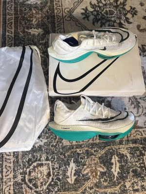 Nike löparskor herr - Nike ZoomX Vaporfly Next% 2 sneakers i vitt med svarta detaljer och turkosgrön sula. Ovandel i mesh för bra andningsförmåga och lätt vikt. Snygg, sportig design med synlig ZoomX-dämpning och klassisk Swoosh-logga på sidan.
