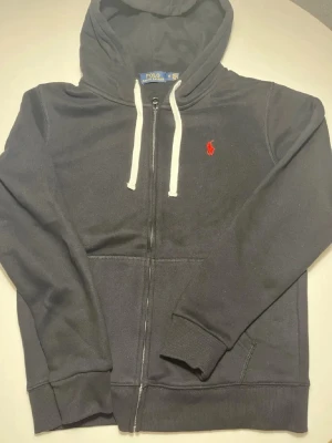 Svart zip hoodie från Polo Ralph Lauren - Svart hoodie med dragkedja från Polo Ralph Lauren. Klassisk design med vit dragsko i huvan och röd broderad logga på bröstet. Tillverkad i mjukt bomullsmaterial och har känguruficka framtill. Perfekt för en avslappnad och stilren look.