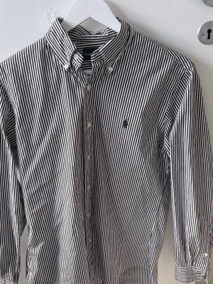 Randig skjorta från Ralph Lauren - Snygg svartvit randig skjorta från Ralph Lauren med klassisk button-down krage och broderad logga på bröstet. Skjortan har långa ärmar och knappar framtill. Perfekt för dig som gillar stilrena plagg med en tidlös vibe.