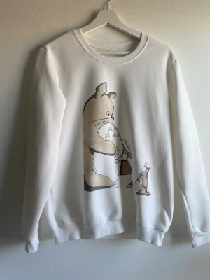 Vit sweatshirt med Totoro-motiv - Vit sweatshirt med tryck av karaktärer från Totoro på framsidan. Tröjan har rund halsringning, långa ärmar och ribbade muddar vid ärmslut och nederkant. Perfekt för dig som gillar anime. OBS, varan har tecken på användning: se bild 2.