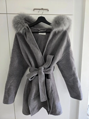 Grå kappa med päls - Stilren grå kappa med stor huva kantad av mjuk päls. Kappan har knytskärp i midjan och två stora fickor framtill. Materialet är en ullblandning. Perfekt för hösten och när det börjar bli varmare efter vintern. Köpt år 2024, Storleken är S/M.