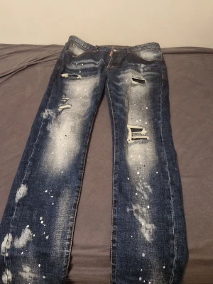 Mörkblå distressed jeans med färgstänk - Säljer ett par mörkblå jeans med slitna detaljer och hål på framsidan samt coola vita färgstänk över hela byxorna. Jeansen har raka ben och klassisk femficksdesign. Perfekt för dig som gillar en edgy och trendig streetstil.