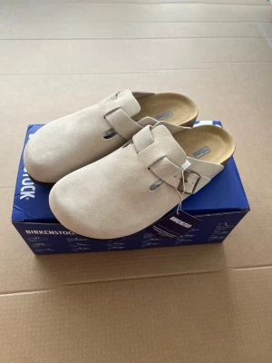 Beige Birkenstock Boston tofflor - Snygga och klassiska Birkenstock Boston tofflor i beige mocka med justerbart spänne i brunt. Tofflorna har en mjuk innersula och en stilren, rundad form som passar perfekt för en avslappnad look. Perfekta för dig som gillar bekväma och trendiga skor.
