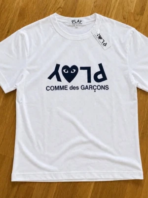 Cdg T-shirt - Snygg vit t-shirt från Comme des Garçons PLAY med det ikoniska hjärtlogotypen och texten 'PLAY' tryckt i mörkblått på bröstet. Klassisk passform med rund hals och korta ärmar. Perfekt för dig som gillar streetwear och stilrena plagg.