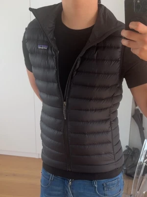 Svart dunväst från Patagonia M - Snygg svart dunväst från Patagonia i storlek M. Västen har quiltad design, hög krage och dragkedja framtill. Är 180cm lång på bilden, skick - 9/10. Nypris är 2 200kr, så ett väldigt bra pris!Kan dock alltid gå ner i pris vid snabb affär 😁🤝. Hör av er vid minsta lilla fundering eller fråga!