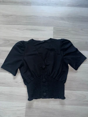 Svart blus med markerad midja - Svart blus med korta puffärmar och markerad smockad midja. Blusen har stora tygklädda knappar framtill och en lätt croppad passform. Perfekt för dig som gillar en trendig och feminin stil.