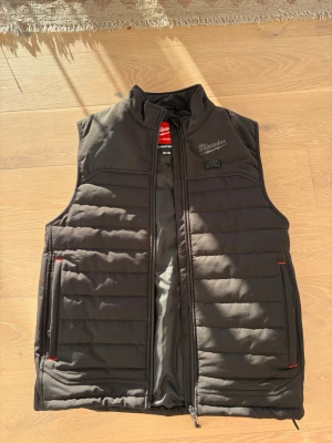 Svart dunväst från Milwaukee S - Snygg svart dunväst från Milwaukee med värmefunktion (M12 Heated Gear). Västen har ståkrage, två dragkedjefickor med röda detaljer och quiltad design. Perfekt för kalla dagar när du vill hålla värmen och ändå vara rörlig. Nedkommer adapter ifall du vill använda m12 batteri.  Jag har Millwaukee powerbank som jag säljer separat. 