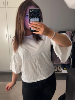Vit blus med volangärmar - Snygg vit blus med vida volangärmar och rund halsringning. Blusen har en rak passform och är tillverkad i ett lätt bomullstyg som känns fräscht och luftigt. Perfekt att styla med jeans eller kjol för en trendig look.