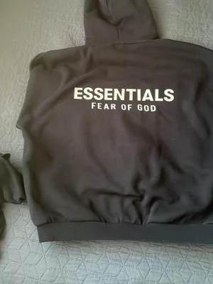 Svart Essentials hoodie Fear of God - Svart hoodie från Fear of God Essentials med vit logotyp på bröstet och ryggen. Klassisk passform med ribbade muddar och huva. Materialet är mjukt och bekvämt, perfekt för en avslappnad streetwear-look.