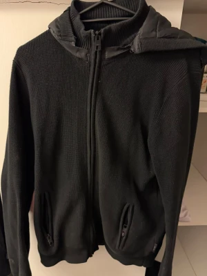 Svart cardigan med huva - Säljer en svart cardigan med dragkedja framtill och två fickor med dragkedja. Tröjan har en quiltad huva i glansigt material och är gjord i ett grovt stickat tyg. Perfekt för dig som gillar en enkel men ändå stilren look.