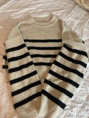 Randig stickad tröja i offwhite och svart - Trendig stickad tröja i offwhite med svarta horisontella ränder. Tröjan har en boxig, croppad passform, ribbad struktur och rund halsringning. Perfekt för lager på lager och ger en cool, avslappnad vibe till din outfit.