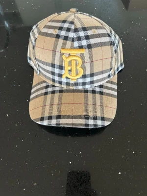 Burberry beige rutig keps - Säljer en klassisk Burberry keps i beige med det ikoniska rutmönstret och ett gult TB-monogram broderat framtill. Kepsen har böjd skärm och justerbart spänne bak i metall. Tillverkad i bomull för en skön och stilren look.